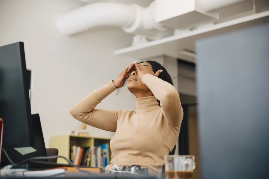 A woman sitting at her office desk leaning back with her hands over her eyes, looking stressed or overwhelmed.