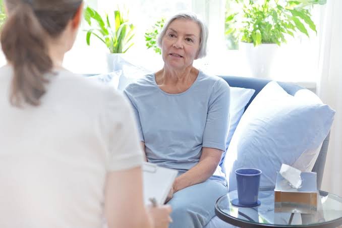 An older woman sitting on a couch speaks with a therapist or counselor holding a clipboard.