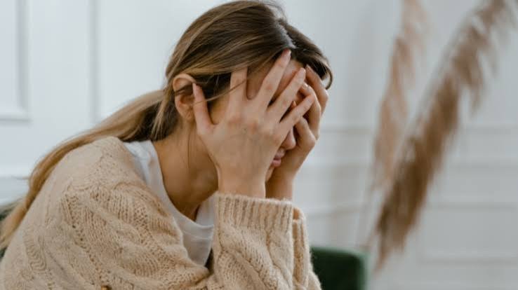 Woman sitting indoors with her face covered by her hands, appearing stressed and overwhelmed.