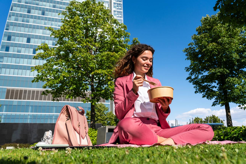 Young woman in a pink suit eating lunch from a takeout bowl on the grass near a modern office building
