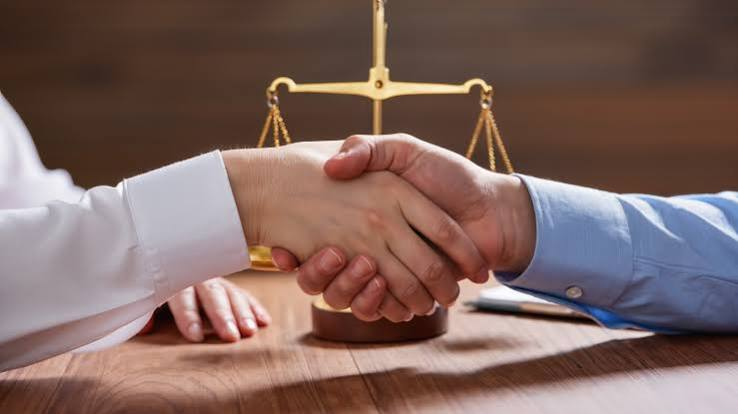 Two people shake hands across a table, with a golden balance scale visible in the background, suggesting a legal or professional agreement.