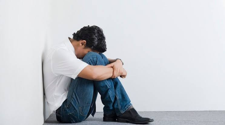 A man sits on the floor against a wall with his knees pulled to his chest and his head lowered onto his arms, appearing distressed or withdrawn.
