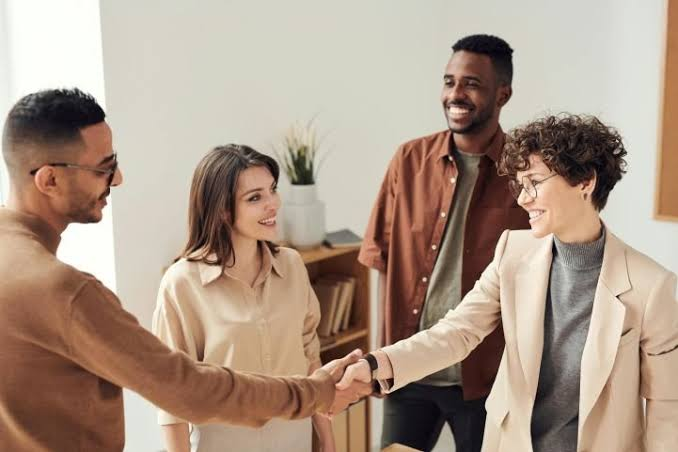 Four coworkers stand together in an office, smiling as two of them shake hands while the others look on.