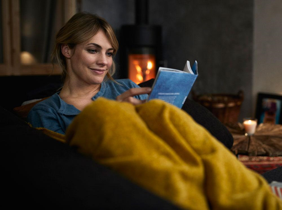 Smiling woman wrapped in a yellow blanket reading a book on the couch by a wood-burning fireplace