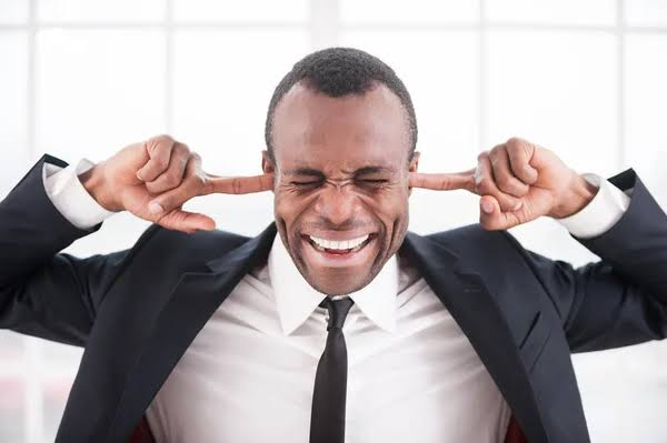 A man in a black suit and tie smiles with his eyes closed while plugging both ears with his fingers.